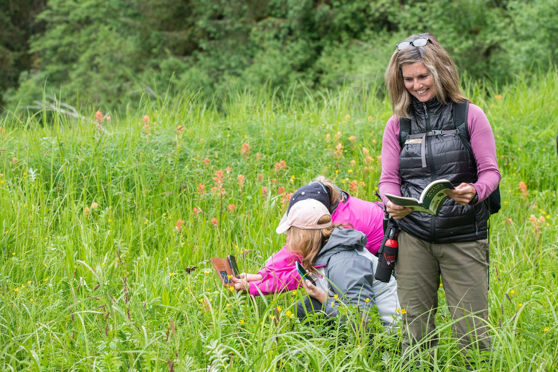 Two guests take a photo of a flower while a naturalist reads a book next to them.