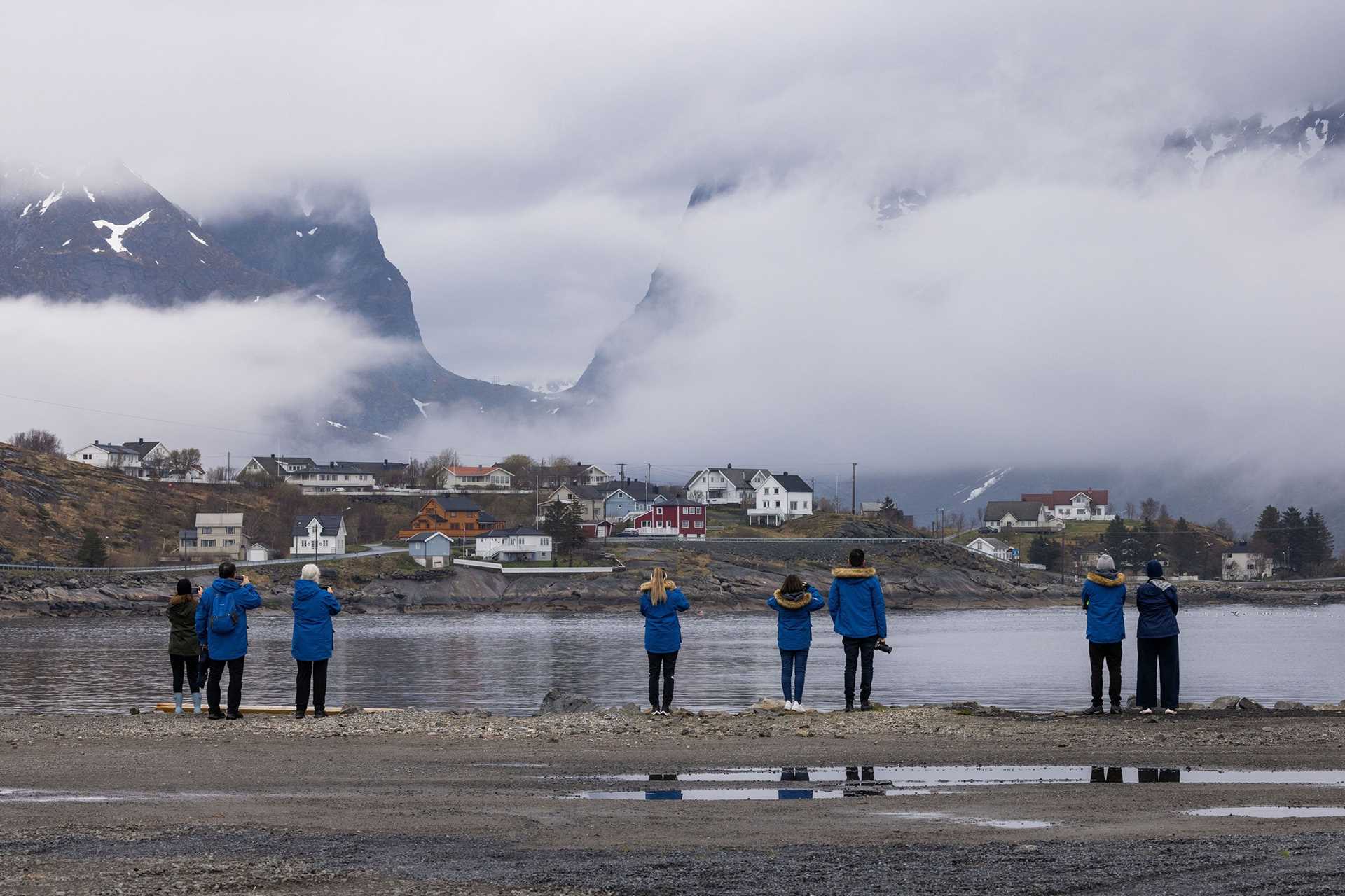 guests in blue parkas take photographs of the town in Reine, Norway