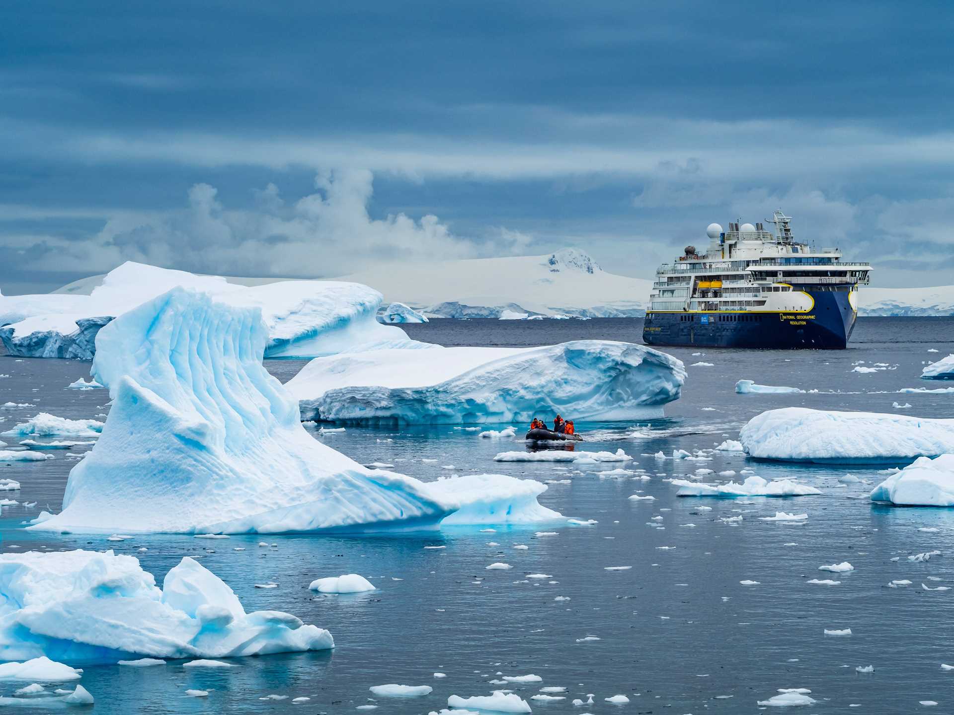 Guests on a Zodiac in the distance cruise through the icebergs in the Gerlache Strait with the National Geographic Resolution in the distance.